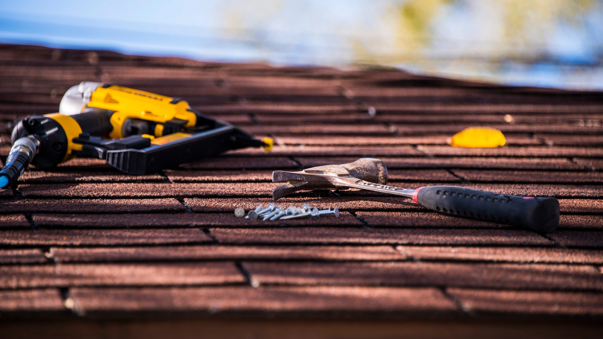 A hammer, nailgun, and roofing nails sitting on a brown shingle roof