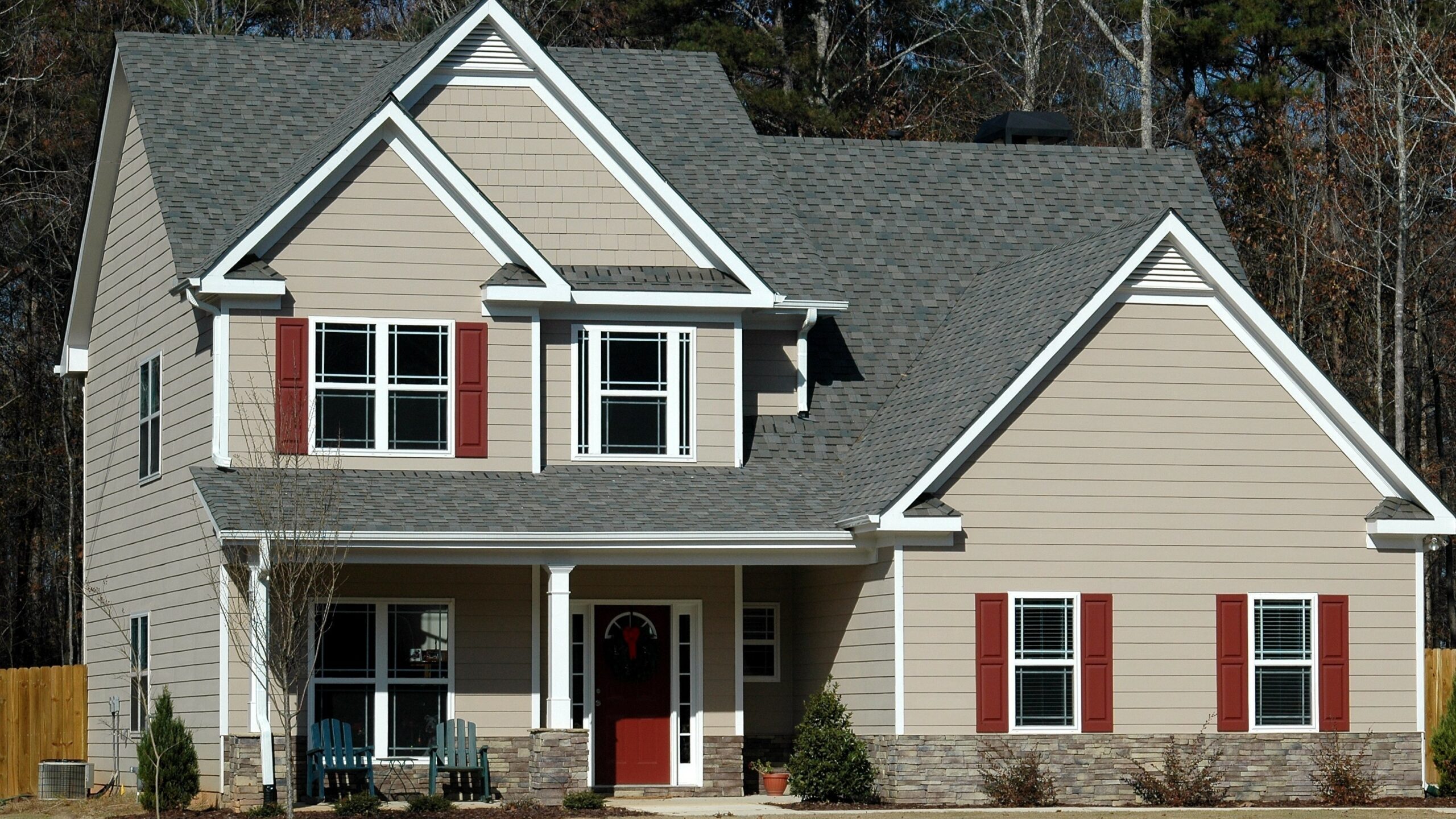 A two-story tan-sided home with white trim, red shutters, and a gray shingle roof