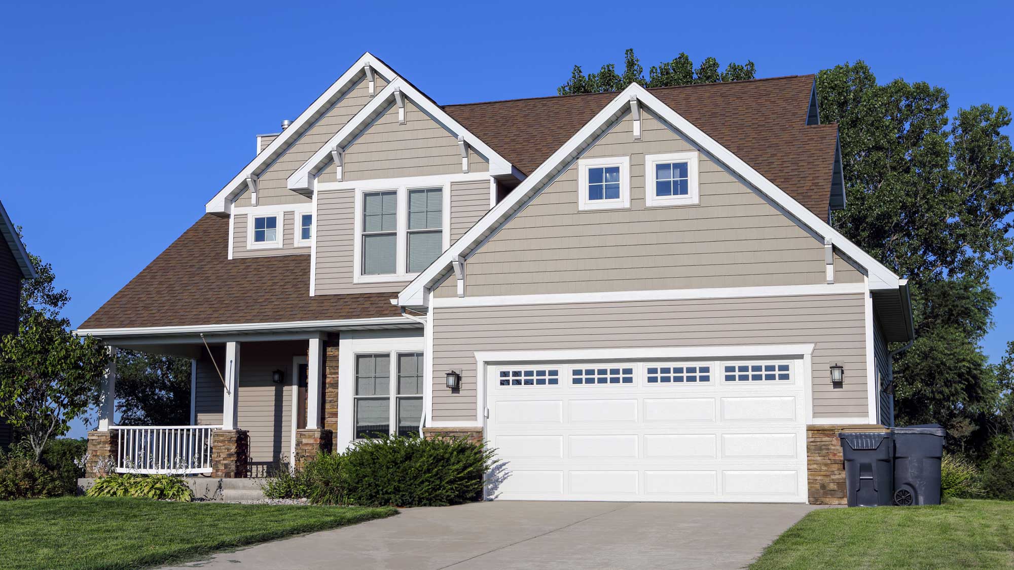 A large two-story home with tan siding, white trim, and a brown asphalt shingle roof