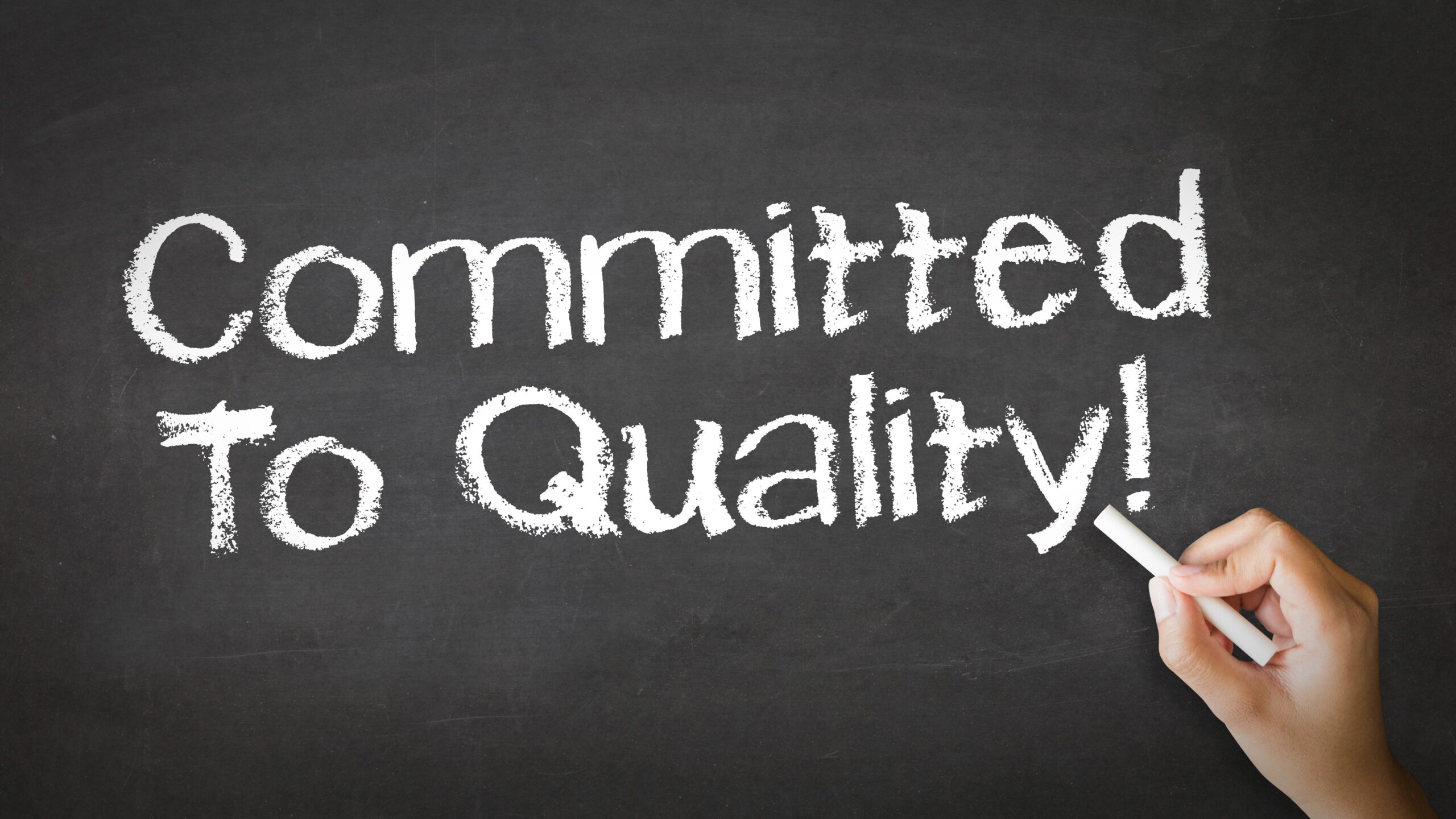 A hand writing a message on a chalkboard that says, “Committed to quality!”